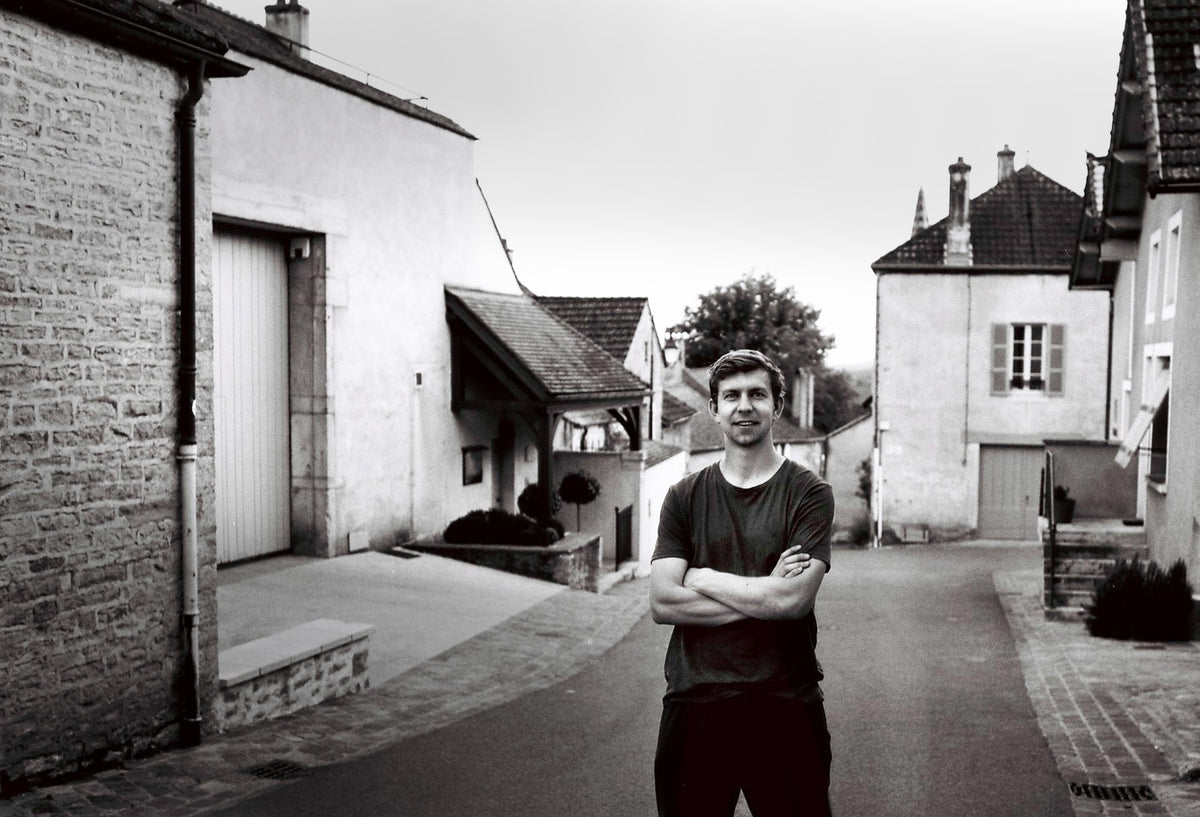 A black and white photograph of Samuel William Roetman Lacey standing with arms crossed in a quiet European village street, framed by historic stone and brick buildings.